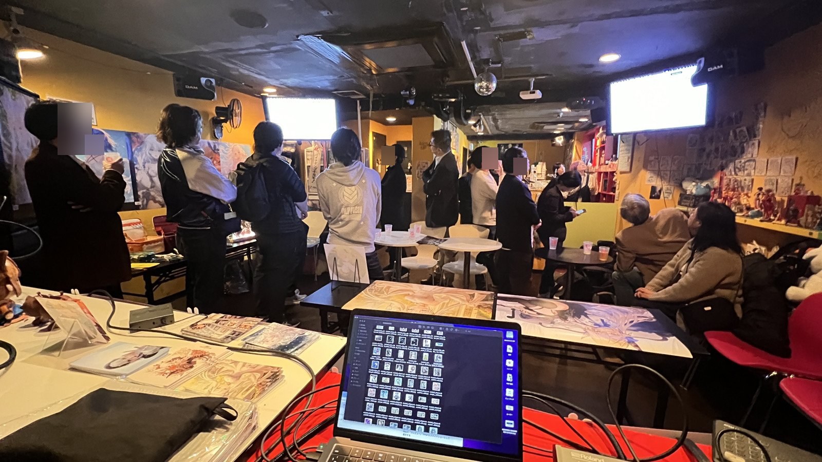 個展会場全景。来場者がモニターと展示物を眺めている薄暗いライブハウス系の会場、手前のテーブルに作品集とノート PC、背景に AI 生成キャラクターポスター複数