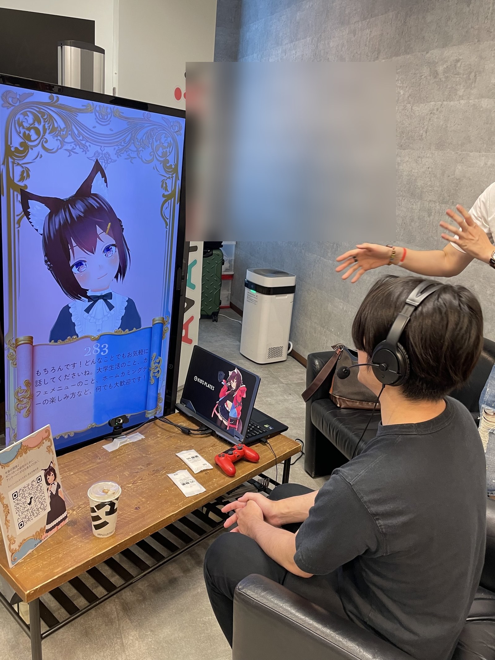 同会場の別アングル。猫耳キャラクターの AI アバター画面と来訪者がヘッドホン経由で対話している様子（来訪者の顔は同意確認不可のためぼかし処理）