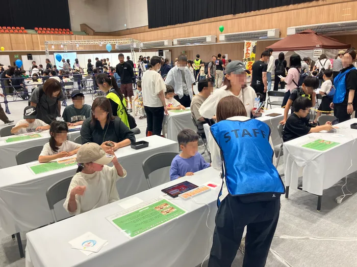 幌延町総合体育館の AI-KATA S2P キッズバージョンのブース。白いテーブルが並び、それぞれの席で来場者がタブレットに向かっている。奥では他の展示に向かう来場者が行き交っている