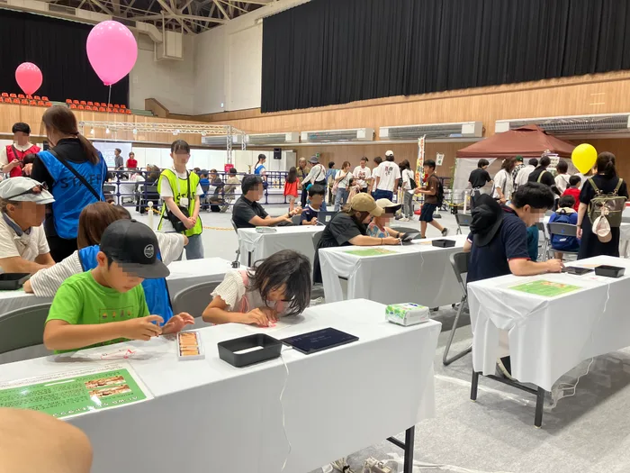 テーブルで画用紙に絵を描く子どもたち。手前には色鉛筆のケースとタブレットが置かれ、天井には来場者が持つピンクの風船が浮かんでいる
