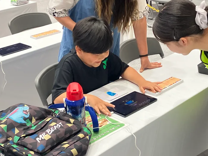 自分で描いた青いキャラクターの絵をテーブルに置き、タブレットを操作する子ども。隣からスタッフが手を添えて操作を案内している