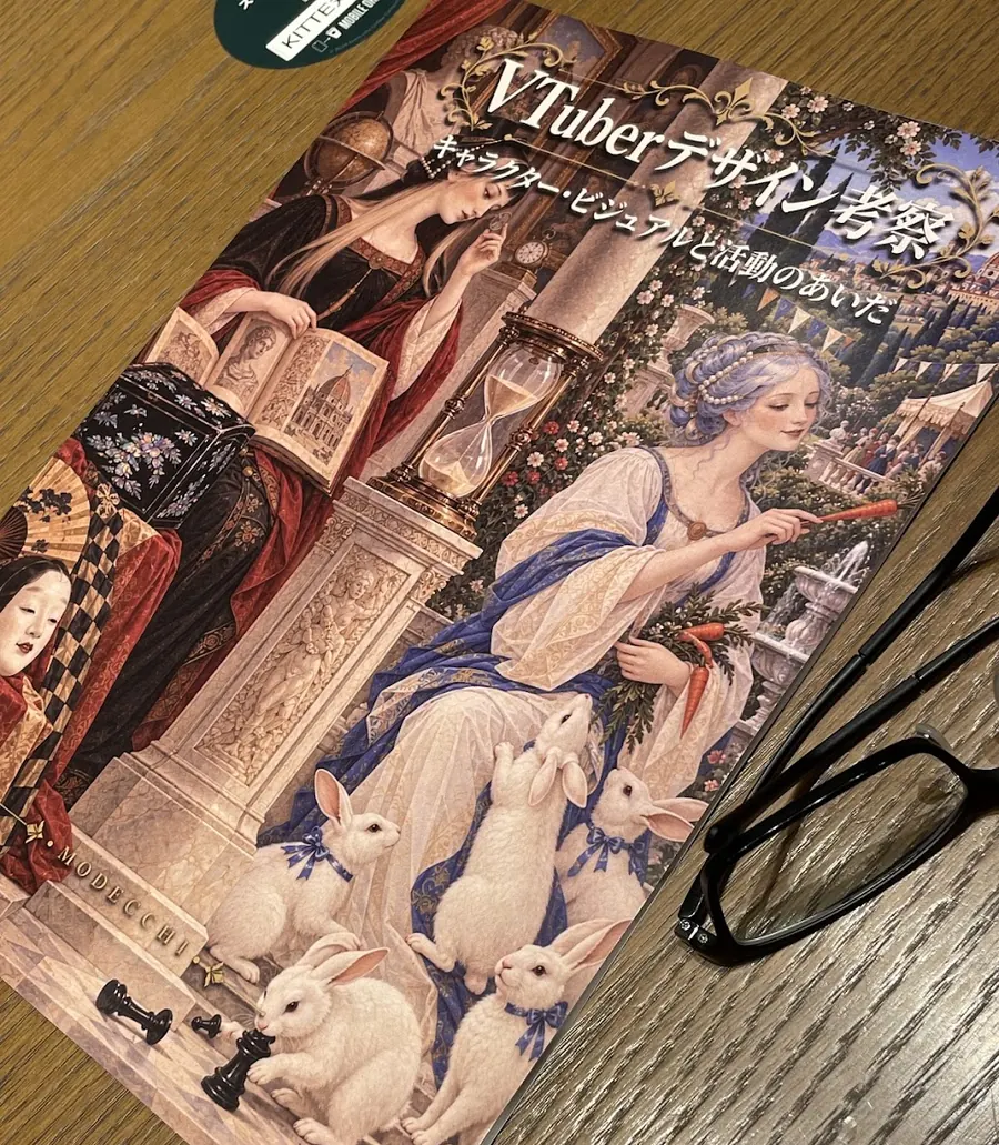 机の上に置かれた同人誌『VTuberデザイン考察　キャラクター・ビジュアルと活動のあいだ』の実物。表紙には砂時計をはさんで書物を読む女性と、にんじんを持って白いウサギに囲まれた女性が古典絵画風に描かれ、左下に MODECCHI の文字がある。右側には眼鏡が置かれている