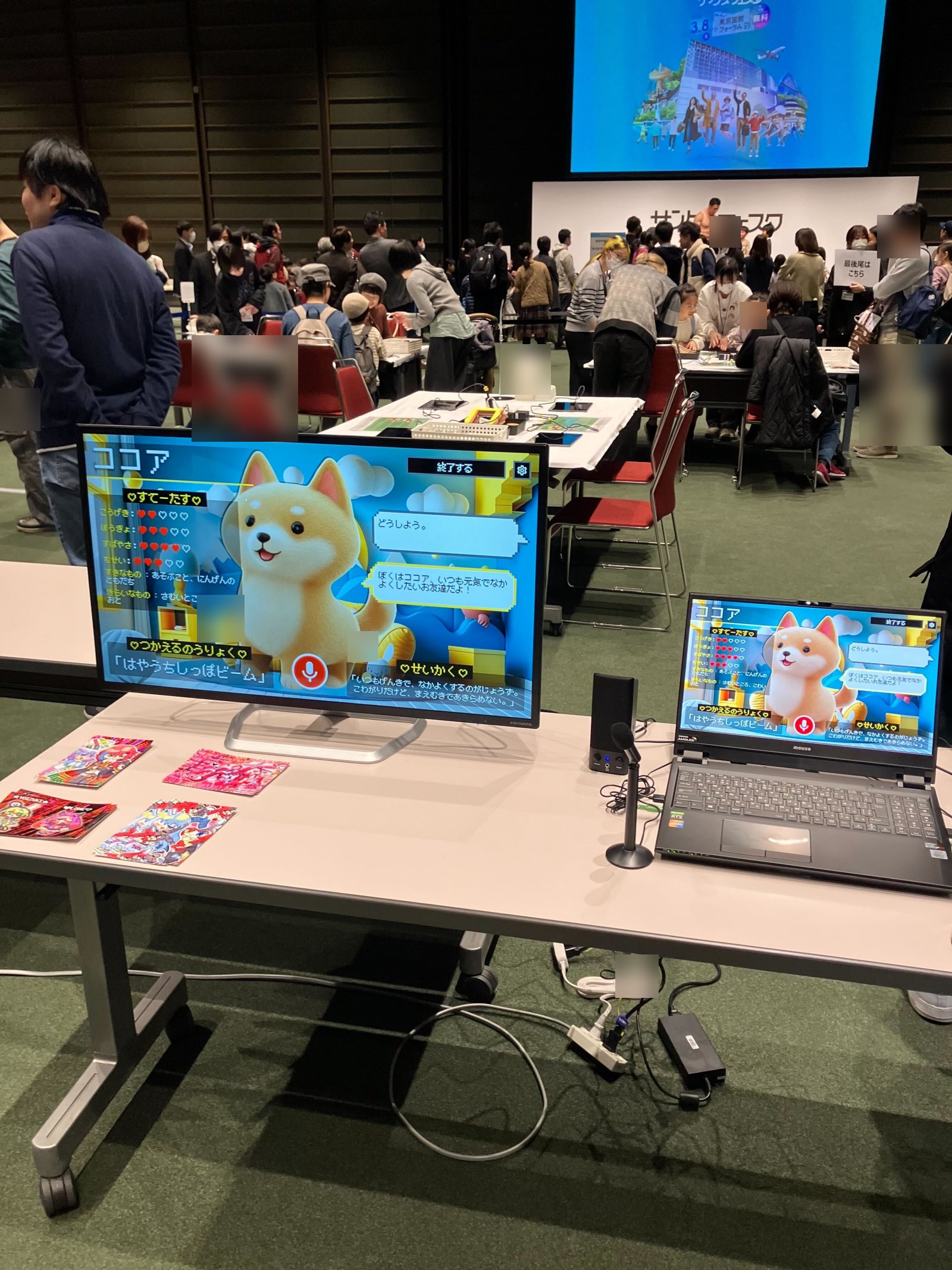ブース全景。大型モニターと PC に AI キャラクター「ココア」、机上に手描き作品カードが並ぶ