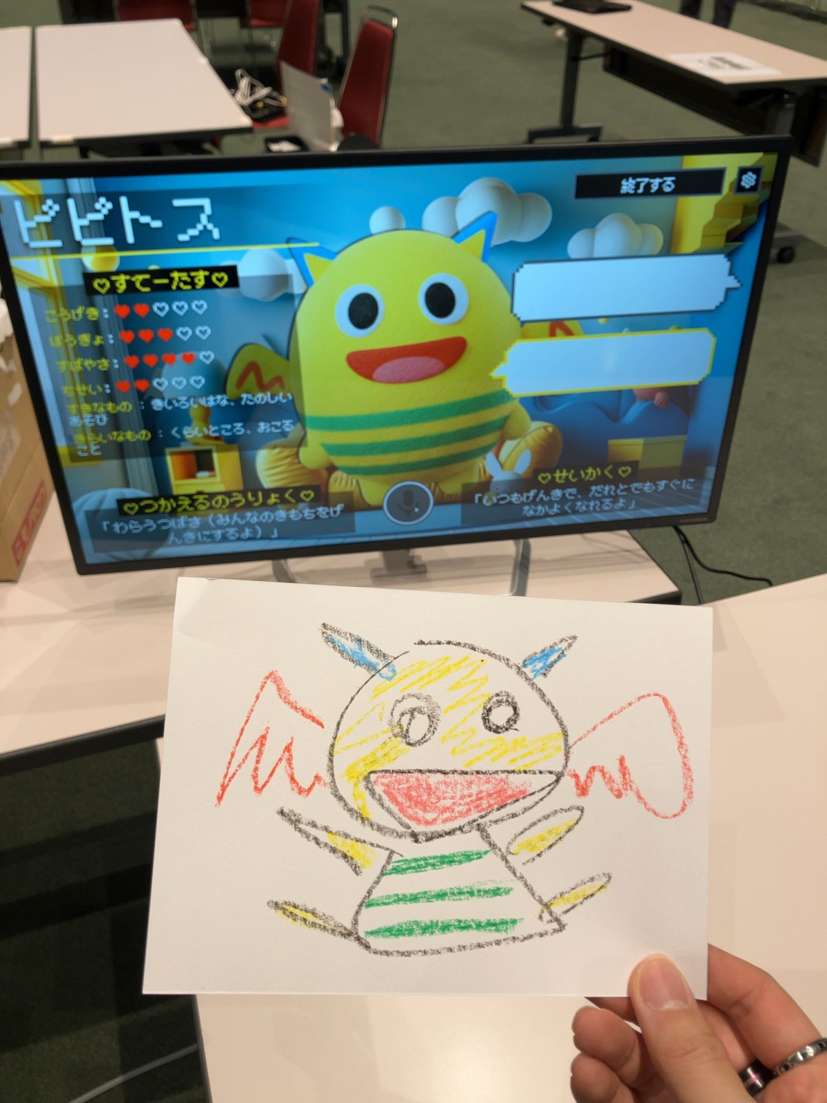 子どもが描いた手描きキャラクター（下、紙）と、AI-KATA S2P キッズが解析・実体化した 3D キャラクター「ビビトース」（上、モニター内）。手描きの黄色いボディと黒い縞、赤い線が、AI 出力では黄色ボディ＋緑シマ＋赤い羽根として変換されている
