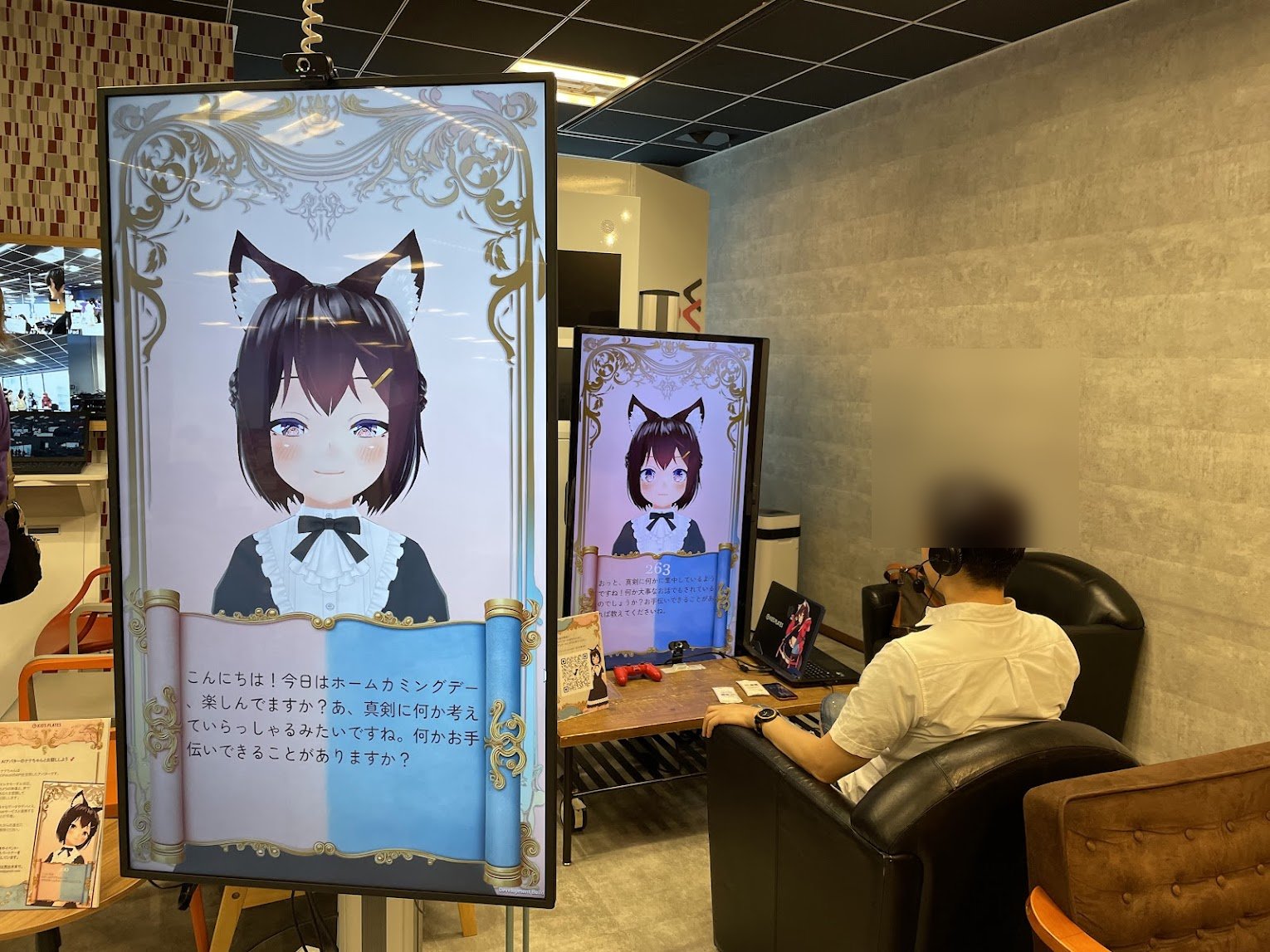 2 画面構成のデモブース。大型縦型モニターと PC モニターに同じ AI キャラクターが表示され、ヘッドホンを着けた来場者が対話している